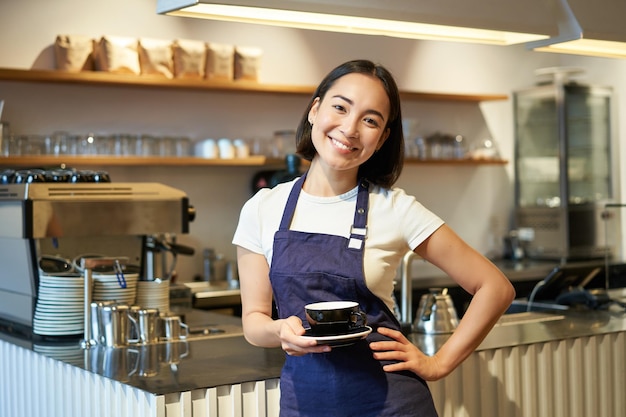 Portrait of smiling asian female barista making coffee holding cup of tea and taking it to cafe clie